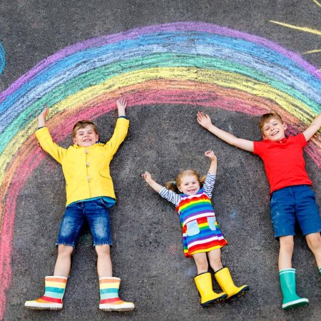 three little children, two school kids boys and toddler girl having fun with with rainbow picture drawing with colorful chalks on asphalt. Siblings in rubber boots painting on ground playing together
