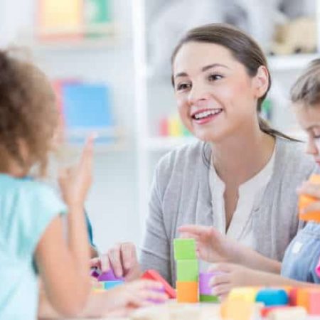 An attentive young kindergarten teacher sits at a table with her students in their classroom.  She looks up from playing with building blocks to listen to an unrecognizable student tell about her weekend.
